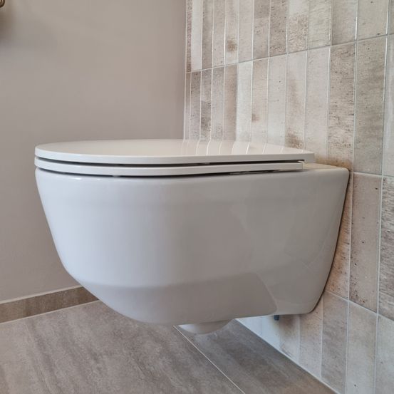 Eine weiße Toilettenschüssel ist an einer Wand mit gefliesten Mustern montiert. Die Wand hat eine hellbeige Farbe und die Fliesen haben eine rechteckige Form mit einer rauen Textur.