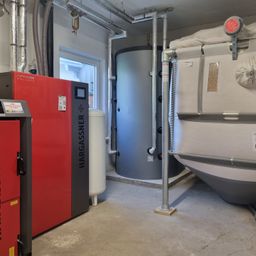 Ein Industrieraum hat eine rote Hargassner-Maschine und einen zylindrischen grauen Wassertank mit angeschlossenen Rohren. In der Ecke steht ein weißer Tank.