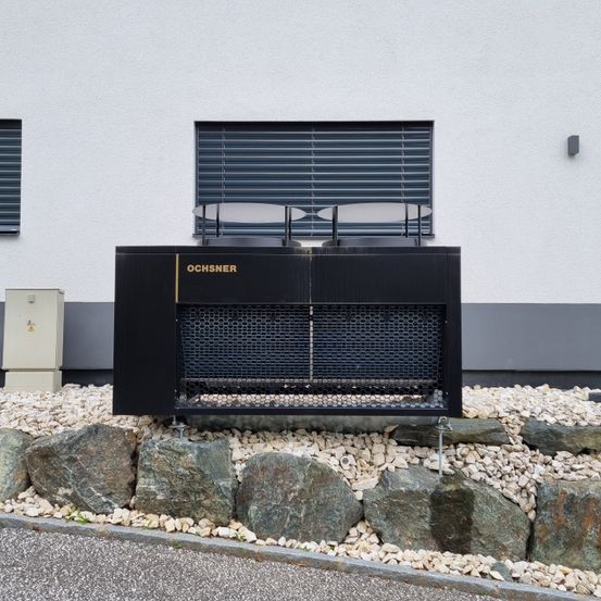 Ein schwarzes Ochsner-Klimagerät steht vor einem weißen Gebäude. Die Einheit ist auf einem Kiesbett platziert, umgeben von Steinen. Über dem Gerät befinden sich zwei Fenster mit Jalousien.