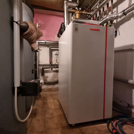 Ein weißes Gerät mit rotem Streifen steht in einem Raum mit gefliestem Boden. In der Nähe befinden sich Metallrohre, eine weiße Wand und ein großer Metallzylinder.