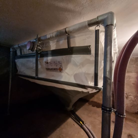 Ein industrieller Tank mit Metallrahmen hängt von der Decke. Er hat eine weiße Abdeckung mit einem Etikett, das 'MAGASSNER' liest. Ein roter Schlauch ist daran angeschlossen.