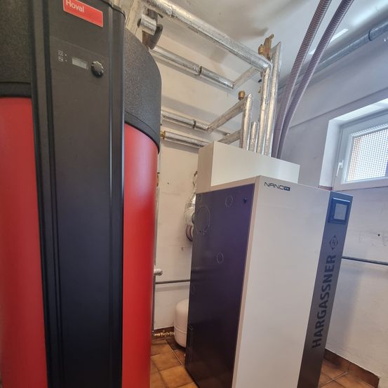 Ein Heizgerät und ein Speichertank sind in einem Raum mit gefliestem Boden und einem Fenster mit Jalousien installiert.