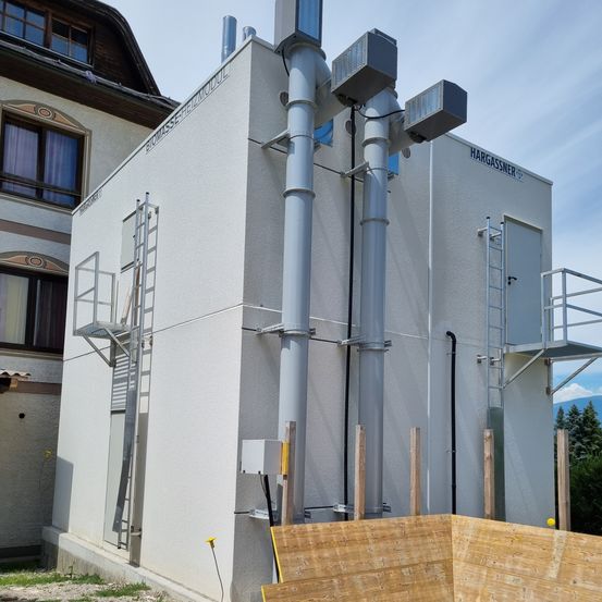 Ein weisses Gebäude mit dem Schild BIOMASSE FERMMODEL und einem HARGASSNER-Label wird gebaut. Es hat eine Leiter auf der linken Seite, Rohre und hölzerne Pfosten davor.