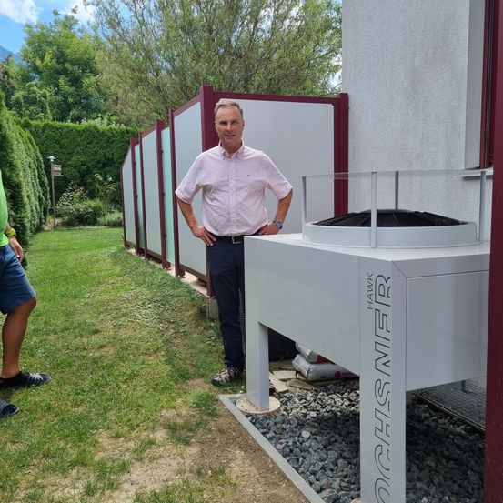 Ein Mann in einem rosa Hemd steht neben einer Glasbox mit der Aufschrift 'OGHS MFR' in einem Hinterhof mit Kiesweg und Garten.