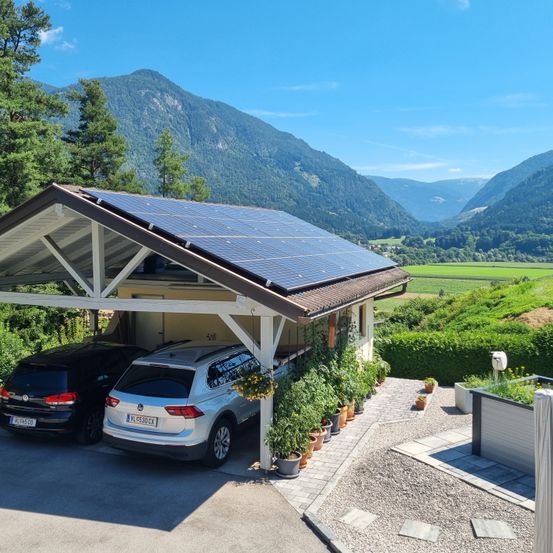 Ein solarbetriebener Carport mit zwei geparkten Autos darunter. Es gibt Bäume, Pflanzen und Solarpanele in der Umgebung. Der Himmel ist blau mit Wolken.