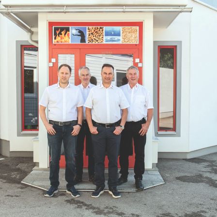 Vier Männer stehen vor einem Gebäude mit roter Tür und Fenster und lächeln für ein Foto. Sie tragen weiße Hemden und schwarze Hosen.
