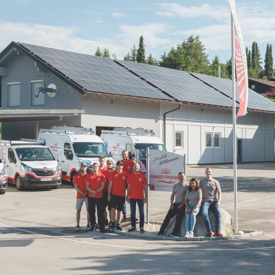 Eine Gruppe von Personen in roten Uniformen posiert vor einem Gebäude mit Solarmodulen. Drei Lieferwagen sind davor geparkt.