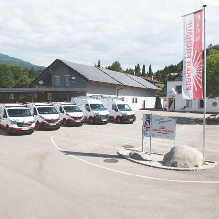 Ein Parkplatz mit mehreren weissen Vans und einer roten Fahne mit Text vor einem Gebäude mit Solarpanelen.
