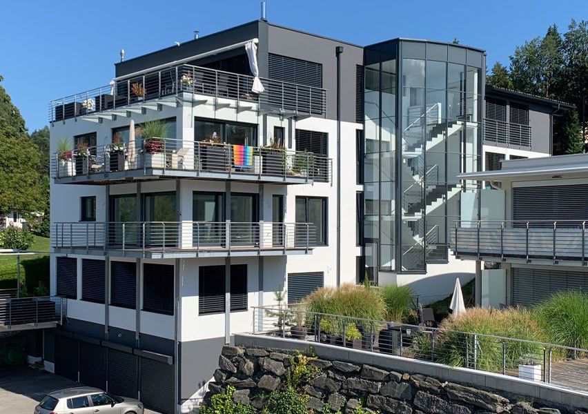Ein modernes mehrstöckiges Wohnhaus mit Glaswänden und Balkonen. Das Gebäude verfügt über einen gläsernen Aufzug, eine Treppe und mehrere Topfpflanzen auf den Balkonen.
