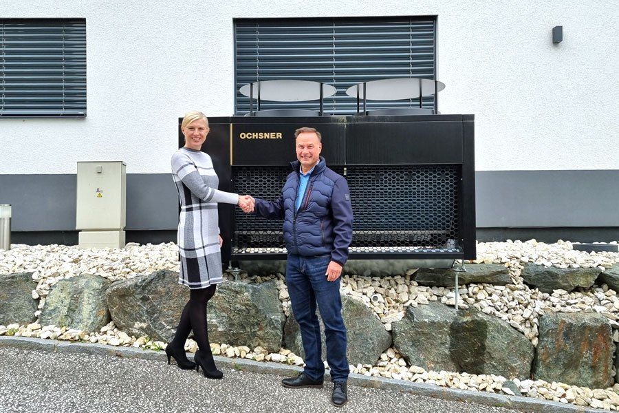 Ein Mann und eine Frau stehen vor einem Gebäude und schütteln Hände. Dahinter steht eine schwarze Maschine mit dem Wort 'OCHSNER' darauf. Sie lächeln beide.