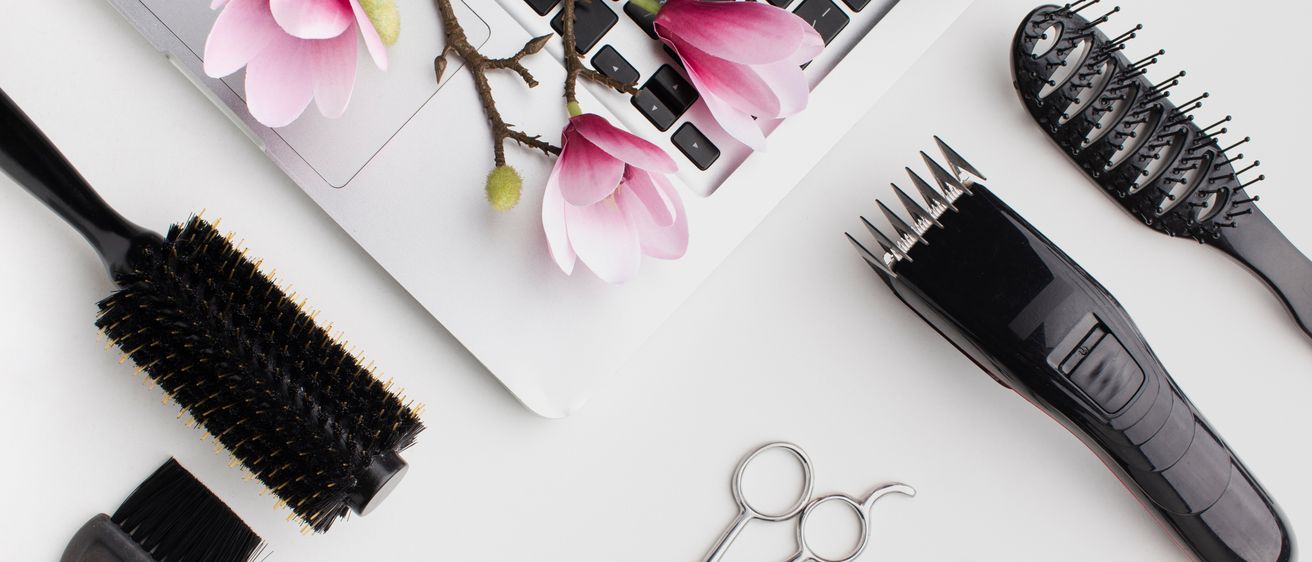 Ein weißer Laptop mit rosa Blumen darauf steht auf einer weißen Oberfläche. Andere Gegenstände sind Scheren, eine Haarbürste und ein Haarschneider.