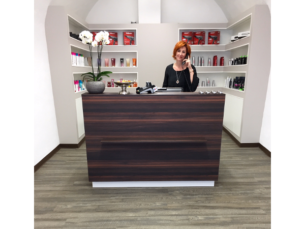 Eine Frau mit schwarzem Oberteil und Halskette telefoniert an einem Rezeptionstresen im Salon. Auf dem Tresen steht eine weiße Blume in einem grauen Topf.