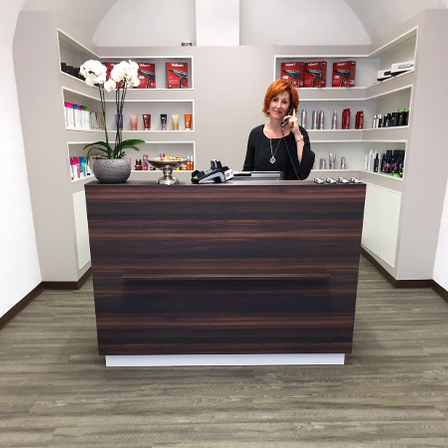 Eine Frau mit schwarzem Oberteil und Halskette telefoniert an einem Rezeptionstresen im Salon. Auf dem Tresen steht eine weiße Blume in einem grauen Topf.