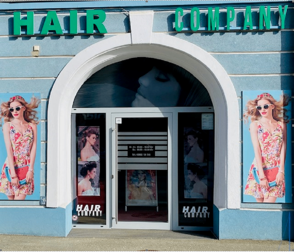 Außenansicht eines Salons namens Hair Company mit einem bogenförmigen Eingang mit Glasscheiben. Auf beiden Seiten des Eingangs befinden sich Werbeanzeigen mit Frauen in Blumenkleidern und Sonnenbrillen.