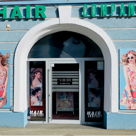 Außenansicht eines Salons namens Hair Company mit einem bogenförmigen Eingang mit Glasscheiben. Auf beiden Seiten des Eingangs befinden sich Werbeanzeigen mit Frauen in Blumenkleidern und Sonnenbrillen.