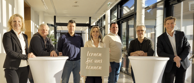 Menschen stehen bei einer Veranstaltung von Bruno Freiluft an einem Podium. Auf dem Podium steht der Text 'Lernen Sie uns kennen'. Die Veranstaltung dreht sich um Transformation und Sanierung.