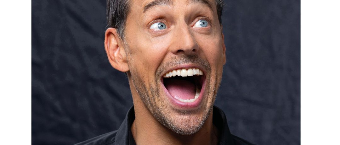 Ein lächelnder Mann mit blauen Augen und Bart, der lacht und den Mund öffnet, trägt ein schwarzes Poloshirt. Der Hintergrund ist dunkel, und der Name Tricky Niki ist unten angezeigt.