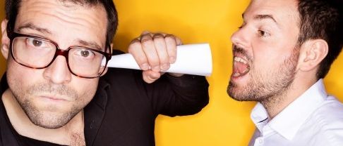 Zwei Männer in einem gelben Raum. Einer mit Brille hält eine Papierrolle, der andere lacht. Unten steht 'FLO & WISC' und darunter 'Musikkabarett: Hörensagen'.