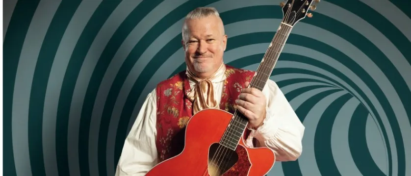 Mike Supancic, in einem roten Weste und weißem Hemd, hält eine rote akustische Gitarre mit einem spiralförmigen Hintergrund. Er lächelt.