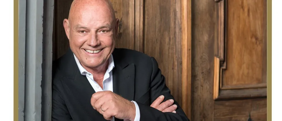 Ein lächelnder Mann in einem schwarzen Anzug steht vor einer Holztür. Er posiert mit verschränkten Armen. Sein Name, Wolfgang Bock, und sein Zitat 'Trautmann Lacht' werden angezeigt.