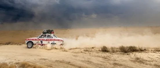 Ein Rallyeauto fährt durch eine Wüste bei bewölktem Himmel. Der Text auf dem Bild lautet 'Kartenpreis 15,-- Tickets im SIB & ONLINE' und 'Rallye Peking - Paris'. Zwei Logos sind in der oberen linken Ecke angezeigt.