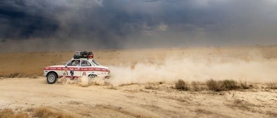 Ein Rallyeauto fährt durch eine Wüste bei bewölktem Himmel. Der Text auf dem Bild lautet 'Kartenpreis 15,-- Tickets im SIB & ONLINE' und 'Rallye Peking - Paris'. Zwei Logos sind in der oberen linken Ecke angezeigt.