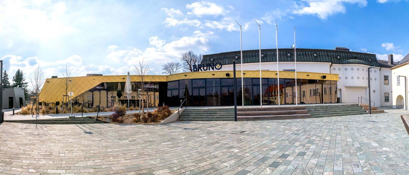Der Eingang des Bruno-Gebäudes mit gelben Wänden, Glasfenstern und Stufen, die unter einem blauen Himmel mit Wolken hinaufführen.