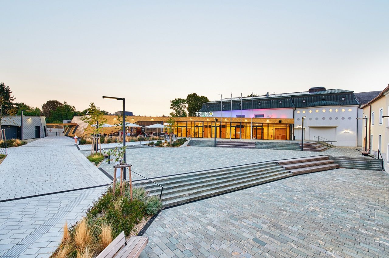 Das Bruno-Theater ist ein modernes Gebäude mit einer goldenen Fassade und Stufen, die zum Eingang führen. Bäume und Pflanzen umgeben es, und eine Bank befindet sich auf der linken Seite.