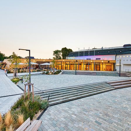 Das Bruno-Theater ist ein modernes Gebäude mit einer goldenen Fassade und Stufen, die zum Eingang führen. Bäume und Pflanzen umgeben es, und eine Bank befindet sich auf der linken Seite.