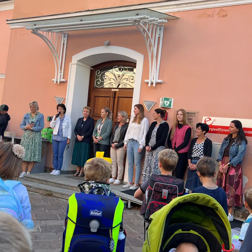 Eine Gruppe von Frauen steht vor einem Gebäude, das von einer Menge beobachtet wird, einige Kinder sind mit Rucksäcken und einem Kinderwagen anwesend.
