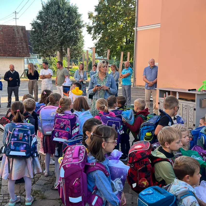 Eine Gruppe von Kindern mit Rucksäcken steht im Kreis, einige Erwachsene stehen hinter ihnen. Sie befinden sich wahrscheinlich bei einer Schulveranstaltung, da im Hintergrund ein Gebäude mit einem großen Fenster zu sehen ist.