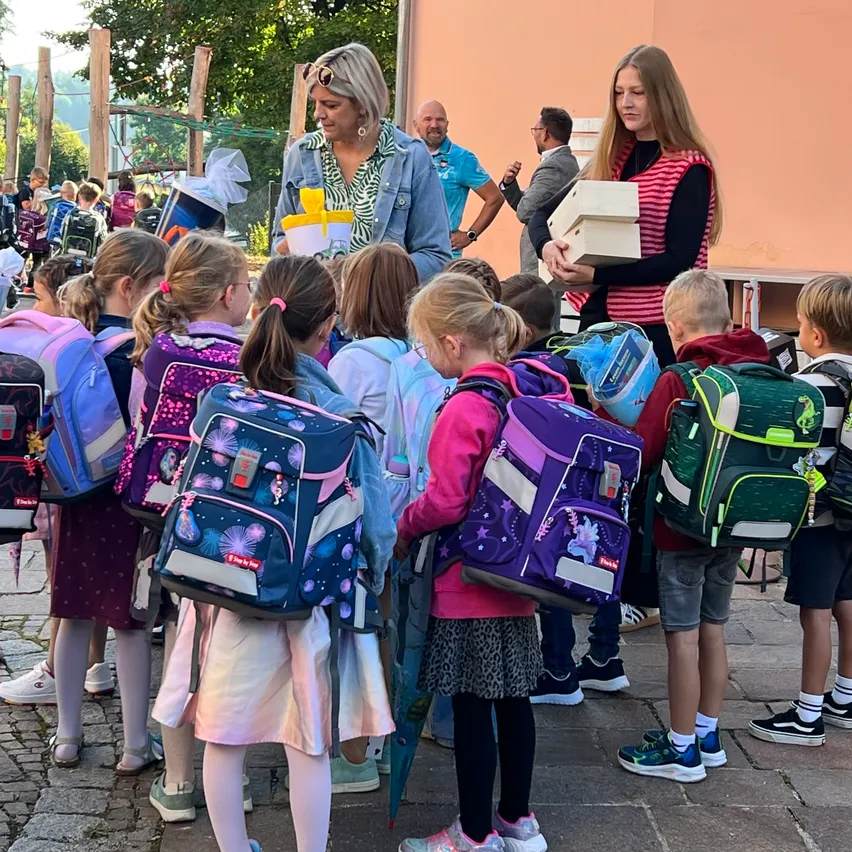 Mehrere Kinder mit Rucksäcken stehen in einer Reihe, wahrscheinlich bei einer Schulveranstaltung. Zwei Frauen, eine mit Brille und einer Jeansjacke, die andere mit einer rosa Weste, halten Schachteln und interagieren mit den Kindern.