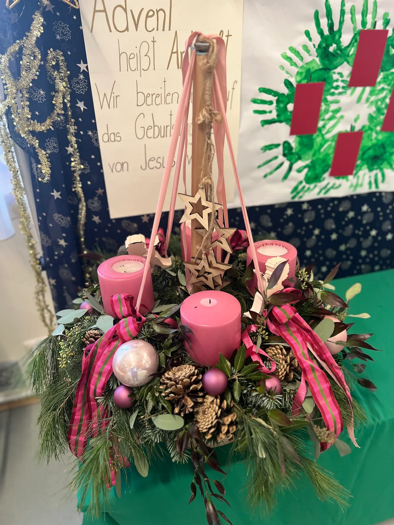 Ein Weihnachtskranz mit vier rosa Kerzen, einer silbernen Kugel und roten Schleifen, geschmückt mit Tannenzapfen und grünen Blättern. Darauf ein Spruch mit Kinderhandabdrücken auf einem Banner.
