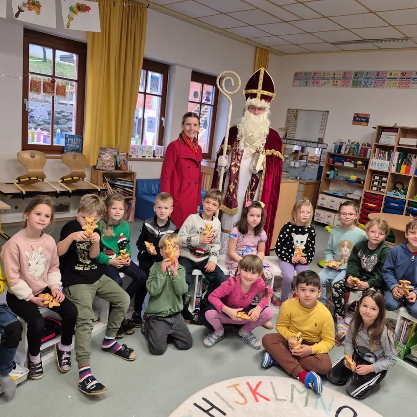 In einem Klassenzimmer sitzen eine Gruppe von Kindern auf dem Boden um den Weihnachtsmann herum und halten Lebkuchen. Dahinter steht eine Frau neben dem Weihnachtsmann. Der Raum ist mit weihnachtlichen Gegenständen dekoriert.