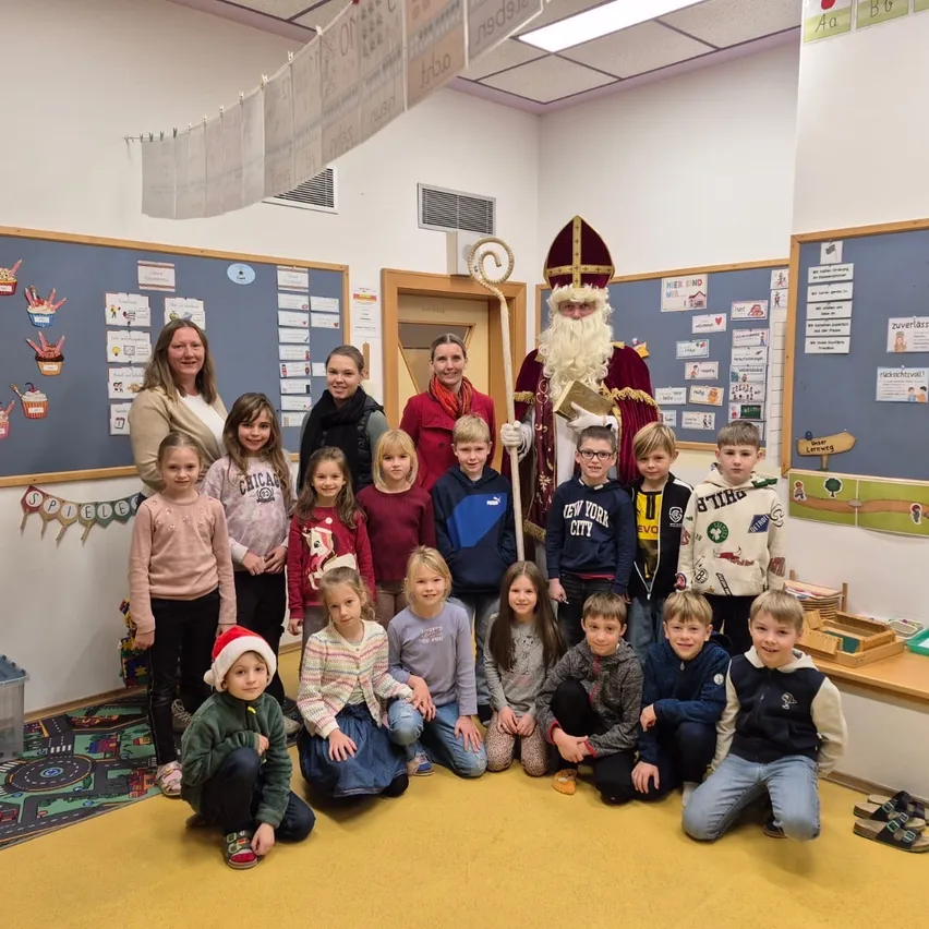 Eine Gruppe von Kindern und Erwachsenen posiert für ein Foto mit einer als Weihnachtsmann verkleideten Person in einem Klassenzimmer.