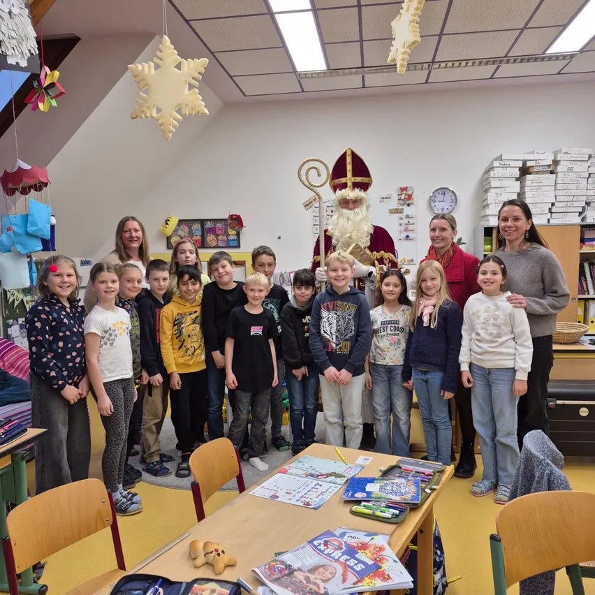 Eine Gruppe von Kindern und Erwachsenen steht in einem Klassenzimmer mit einer Weihnachtsmann-Figur. Ein Schreibtisch mit Büchern, Stiften und Bleistiften steht vor ihnen. Schneeflocken und eine Uhr hängen an der Wand.
