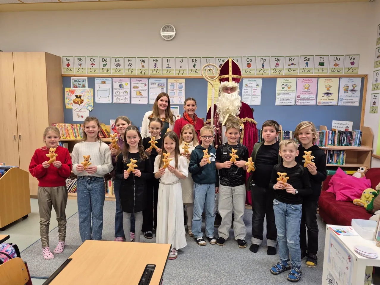 Eine Gruppe von Kindern mit dem Weihnachtsmann, der Kekse in einem Klassenzimmer hält, mit einer Frau und zwei Erwachsenen hinter ihnen. Das Klassenzimmer hat Bücherregale, Poster und eine Uhr.