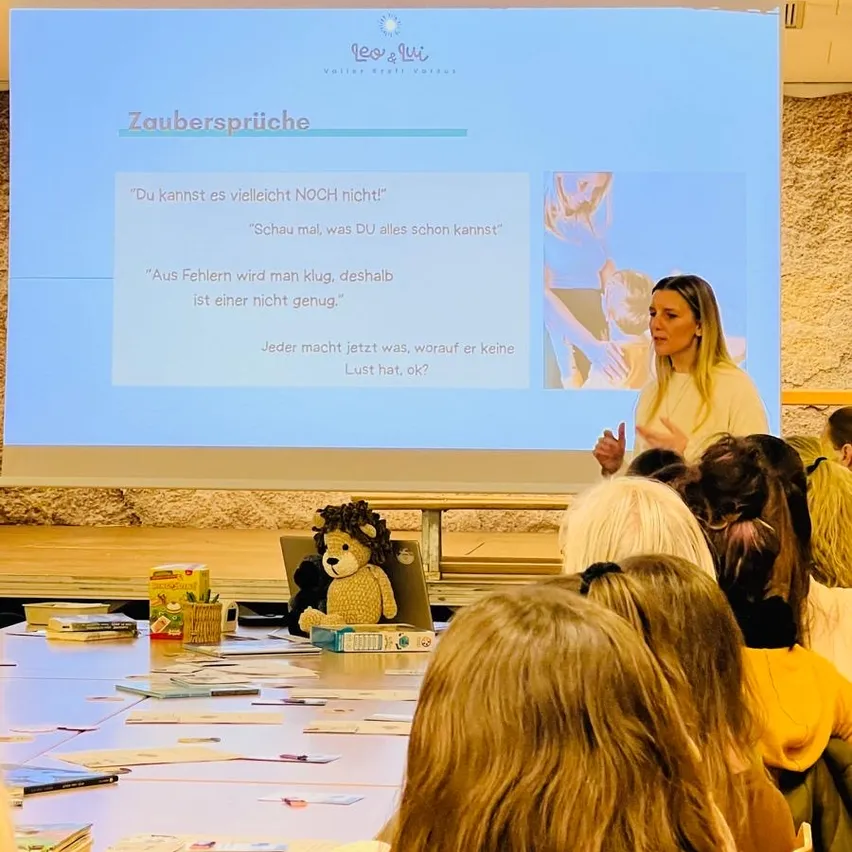 Eine Frau steht vor einem Klassenzimmer und präsentiert eine Folie mit 'Zaubersprüche'. Die Folie enthält Bilder und deutschen Text. Kinder sitzen um einen Tisch mit verschiedenen Gegenständen.