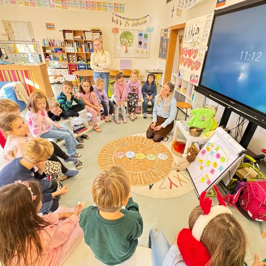 Ein Klassenzimmer mit Kindern im Kreis sitzend, eine Lehrerin spricht. Ein großer Bildschirm zeigt die Zeit 11:12. Der Raum ist mit pädagogischen Postern und Bücherregalen dekoriert.