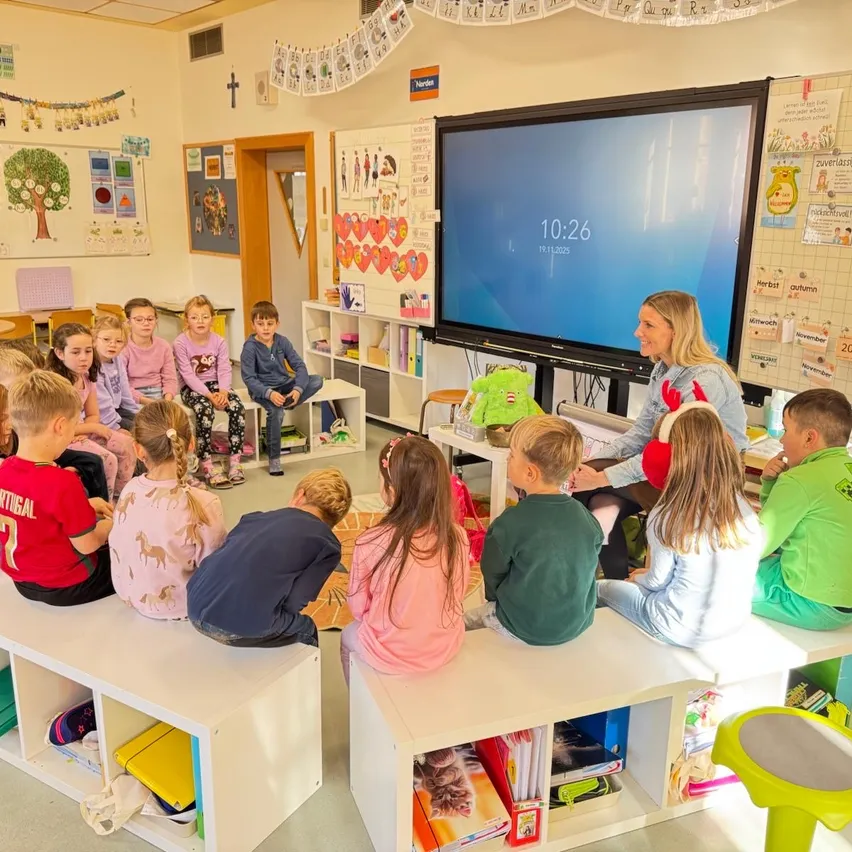 Eine Klassenzimmerszene mit Kindern, die um eine Frau herumsitzen. Die Frau spricht mit ihnen, während ein Monitor hinter ihr die Zeit 10:26 anzeigt. Verschiedene Dekorationen und Gegenstände schmücken die Wände und Regale.