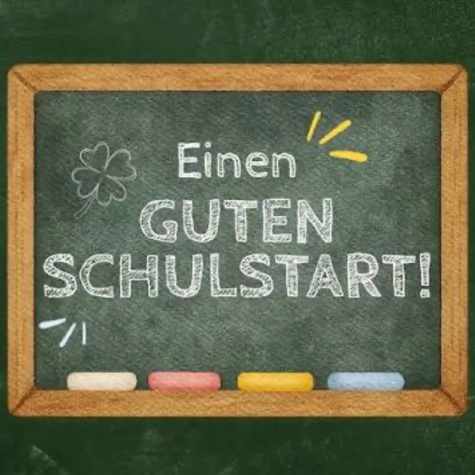 Ein Schultafel mit der Aufschrift 'Einen Guten Schulstart!' in weißer Kreide geschrieben. Ein gelber Sonnenstrahl und ein grünes Kleeblatt sind ebenfalls gezeichnet. Unten befinden sich vier farbige Kreiden.