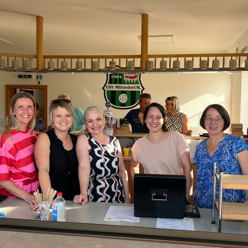 Five women are smiling and posing for a photo at a bar. Behind them is a counter with a monitor, cups, and bottles. There is a logo on the wall.