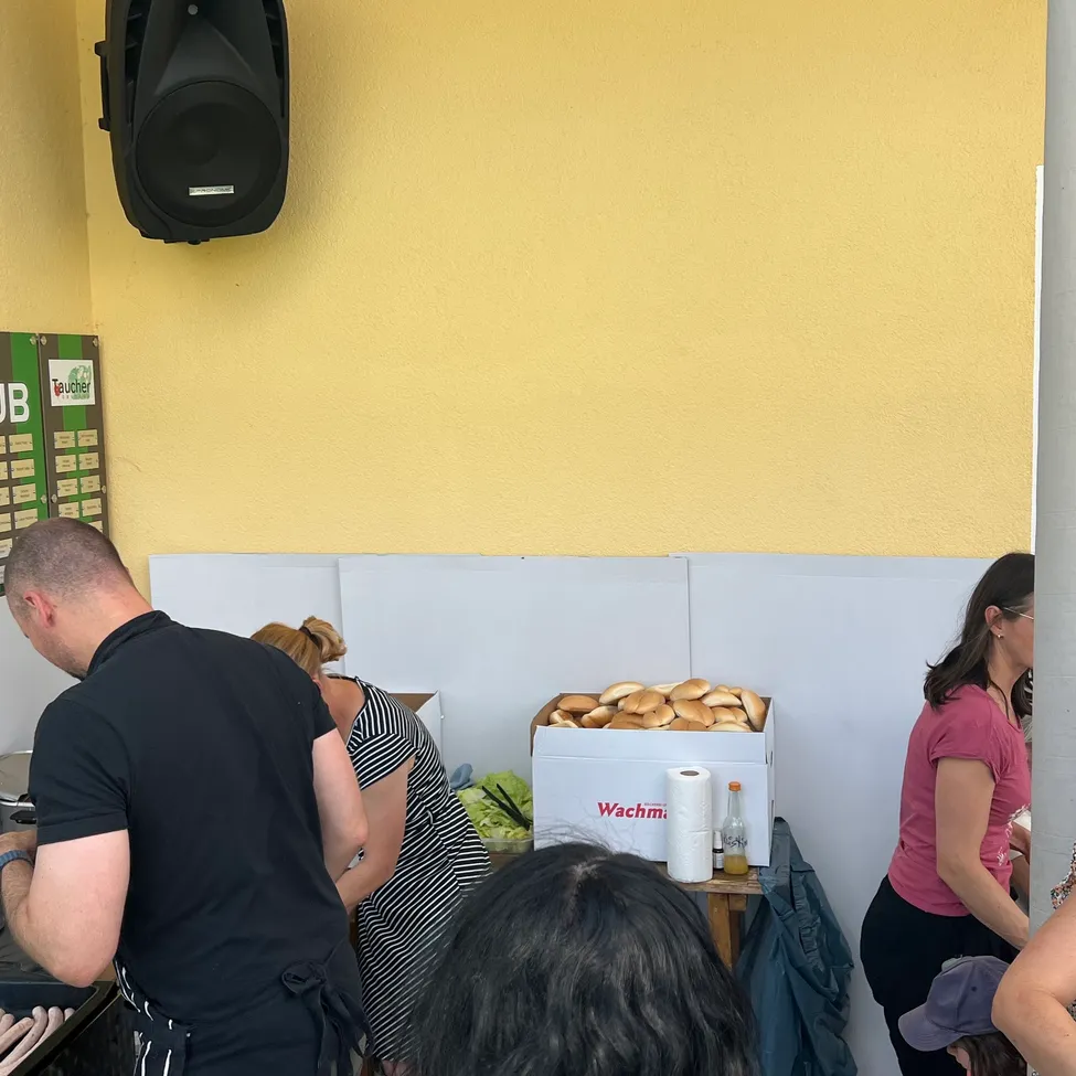 Zwei Personen bereiten an einem Tresen in einem Raum Essen zu. Eine Schachtel Brot steht im Hintergrund auf einem Tisch.