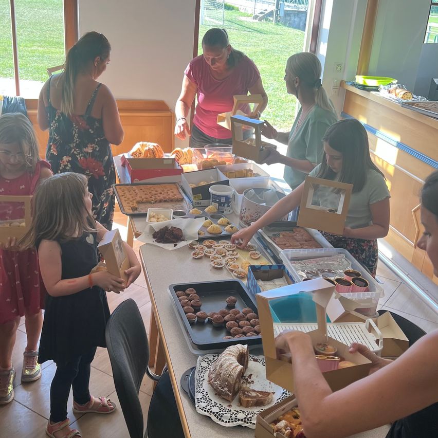 A group of women and girls are gathered around a table filled with various desserts, including pastries and cakes. They are holding boxes and appear to be sorting or selecting items.