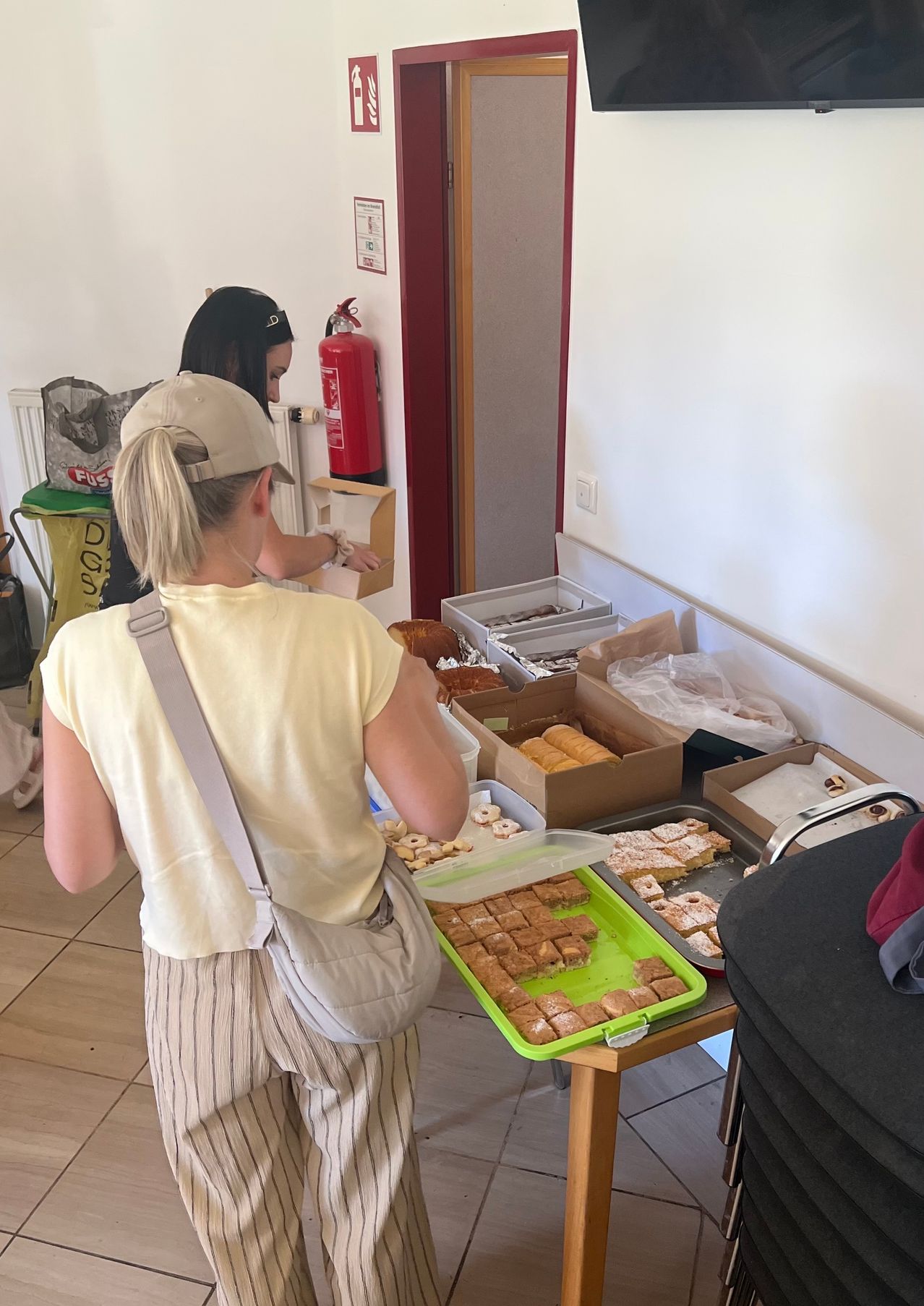 Two women are looking at a table of pastries. One woman wears a yellow shirt and a beige bag. A fire extinguisher is mounted on the wall.