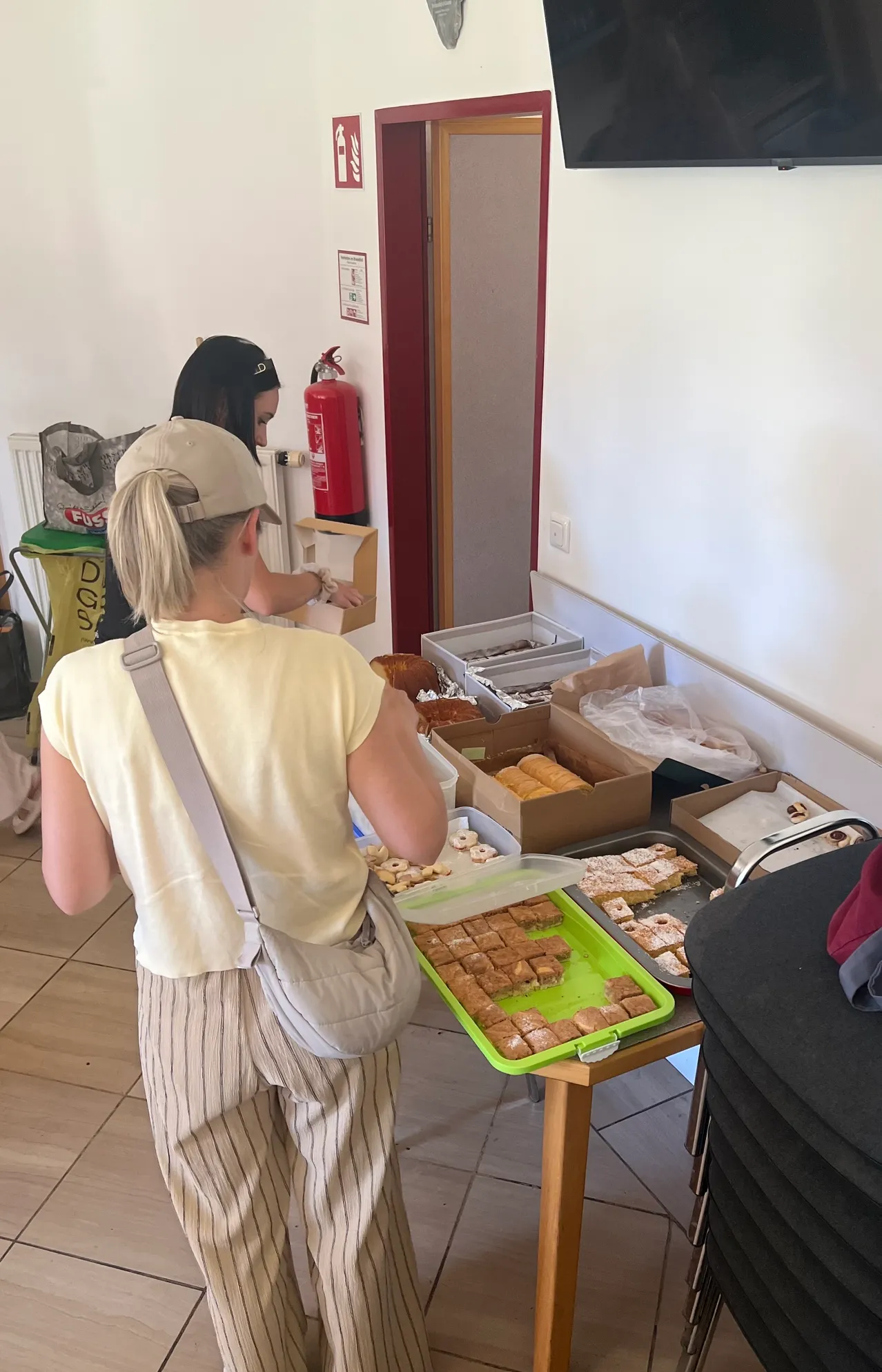 Zwei Frauen betrachten einen Tisch voller Gebäck. Eine Frau trägt ein gelbes T-Shirt und eine beige Tasche. Ein Feuerlöscher ist an der Wand montiert.