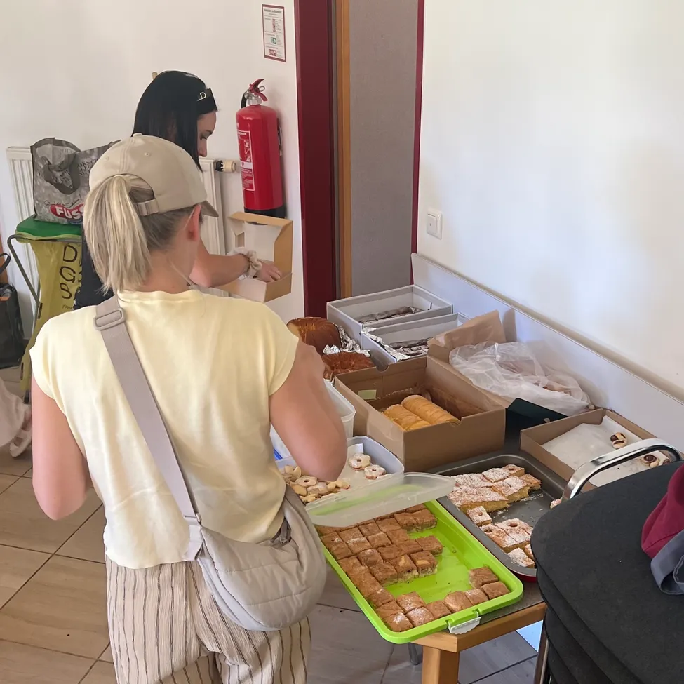 Zwei Frauen betrachten einen Tisch voller Gebäck. Eine Frau trägt ein gelbes T-Shirt und eine beige Tasche. Ein Feuerlöscher ist an der Wand montiert.