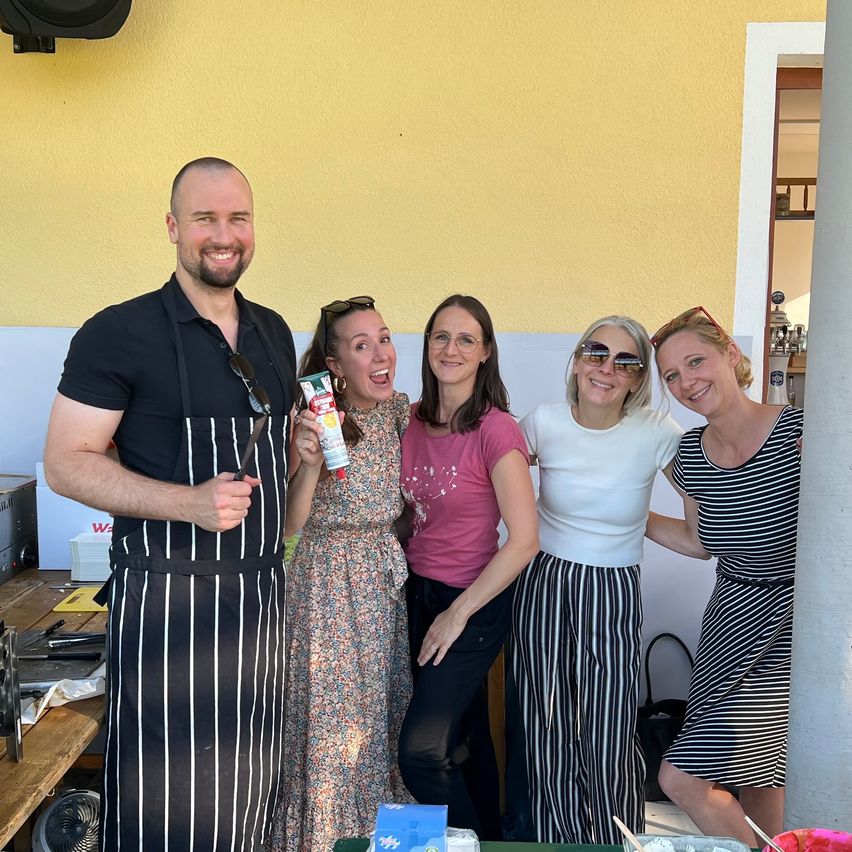 Five smiling people stand close together in a kitchen, a man with a chef's apron holds a bottle. Behind them is a yellow wall, a table with a fan, and a white door.