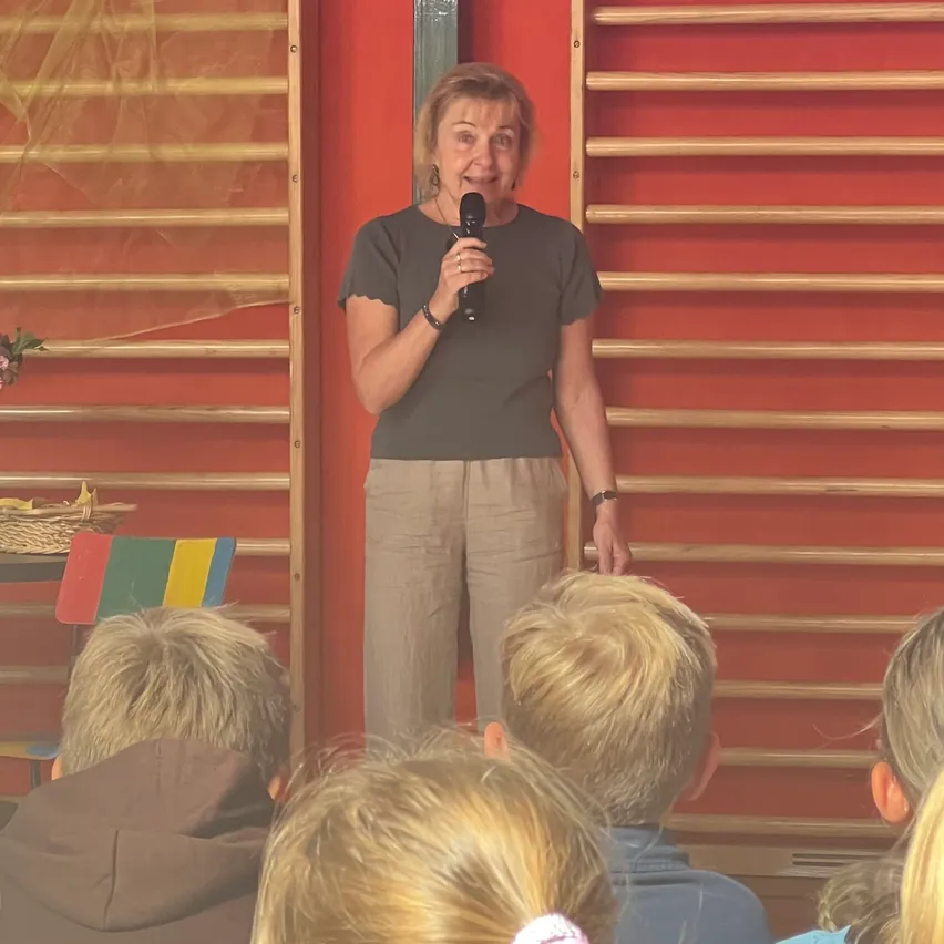 Eine Frau steht vor einer roten Wand, hält ein Mikrofon in der Hand und spricht zu einer Gruppe von Kindern.