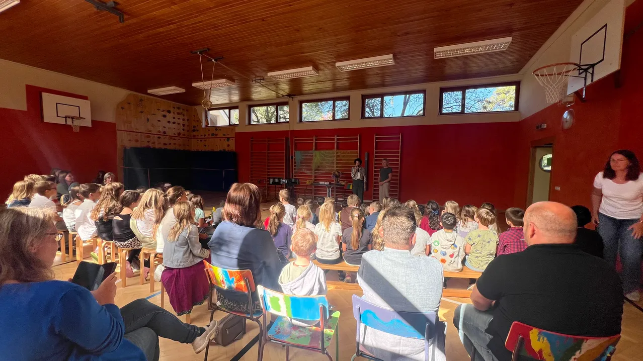 Eine Gruppe von Menschen sitzt in einem Raum mit roten Wänden und einer Holzdecke. Einige sitzen auf Stühlen, andere stehen. Zwei Personen stehen auf der Bühne und eine Frau steht in der Nähe der Tür.